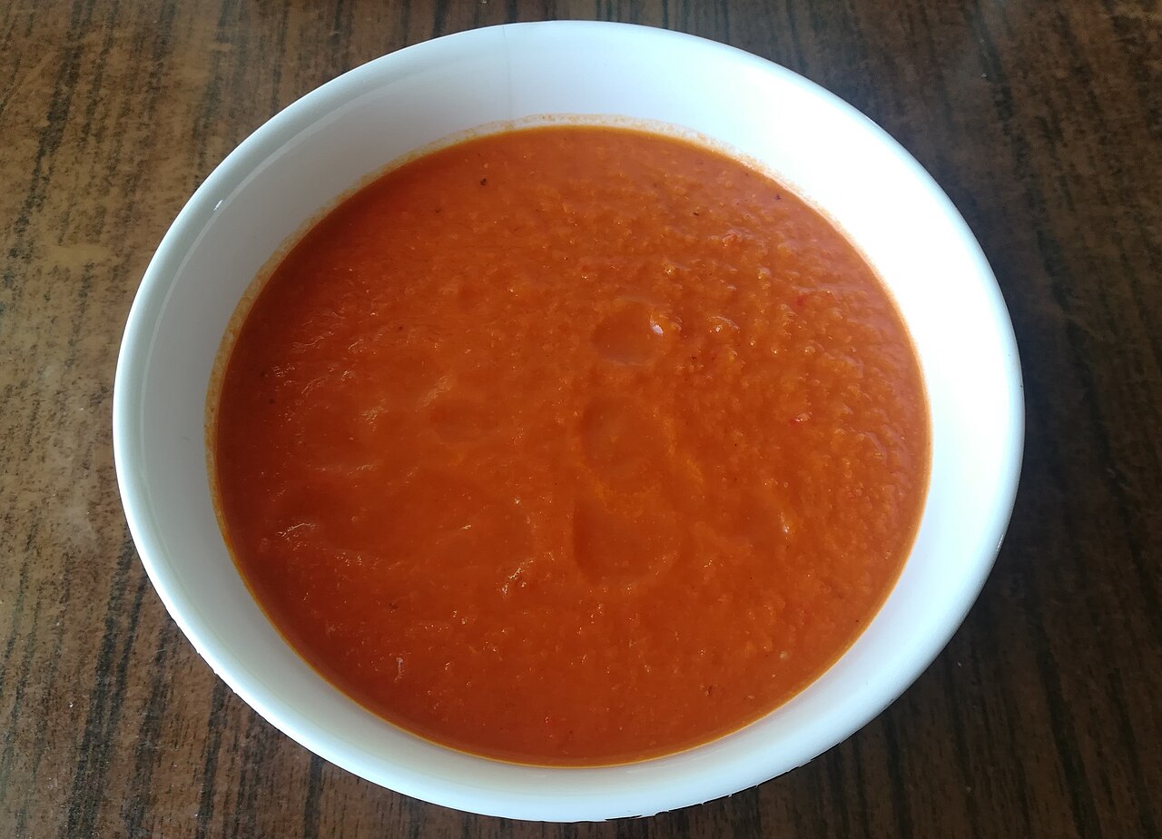 Élénkpiros sült paprika krémleves fehér tálban, fa asztalon