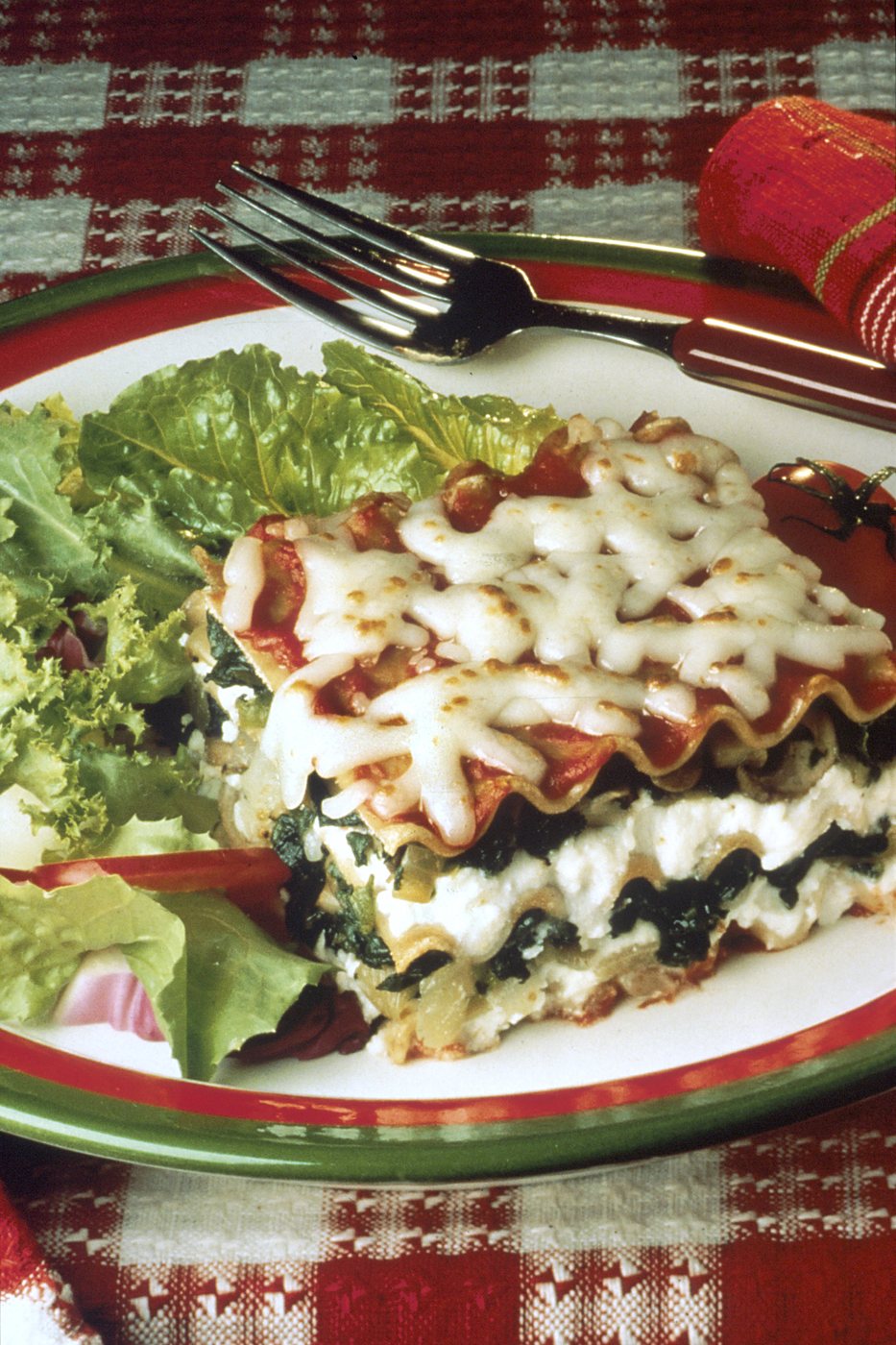 Egy szelet spenótos lasagne tányéron, olvadt sajttal a tetején, zöldsalátával, kockás terítőn